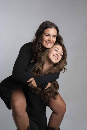 Photo amusante de deux amies en studio, l'une portant l'autre sur son dos, toutes deux souriantes et habillées en noir