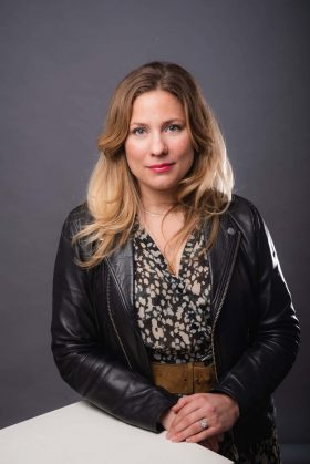 Femme aux cheveux blonds en veste en cuir appuyée sur un support blanc, regardant directement l'objectif avec un léger sourire