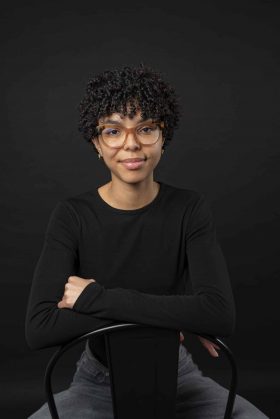 Portrait studio d'une jeune femme à lunettes et cheveux bouclés, assise de manière décontractée sur une chaise, vêtue d'un haut noir