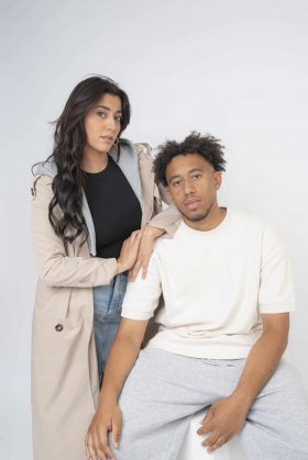 Couple posant en studio avec un style vestimentaire moderne, l'homme assis en blanc et la femme debout en trench beige, regardant l'objectif