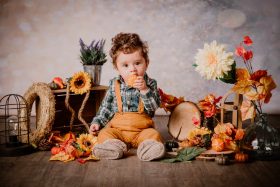 Petit garçon en salopette mangeant une petite citrouille dans un décor de studio automnal avec tournesols et feuilles