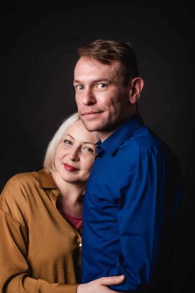Portrait studio couleur d'un couple souriant, l'homme en chemise bleue et la femme en chemise moutarde posant sur fond noir