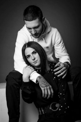 Pose artistique de couple en noir et blanc, la femme assise entre les jambes de l'homme qui l'entoure de ses bras, regard direct